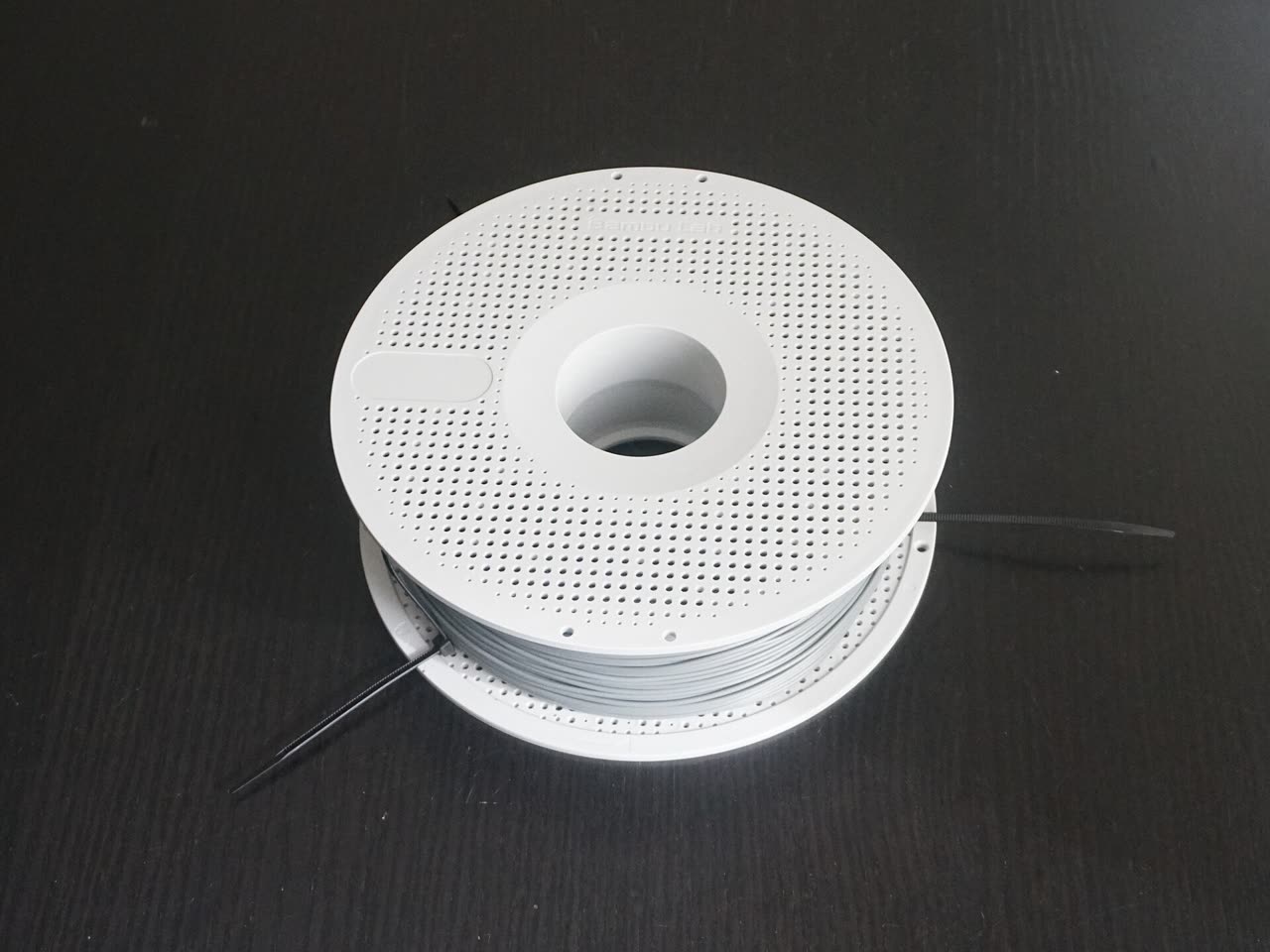 A closed white Bambu Lab spool wound with grey filament, with black zip ties still on the roll.