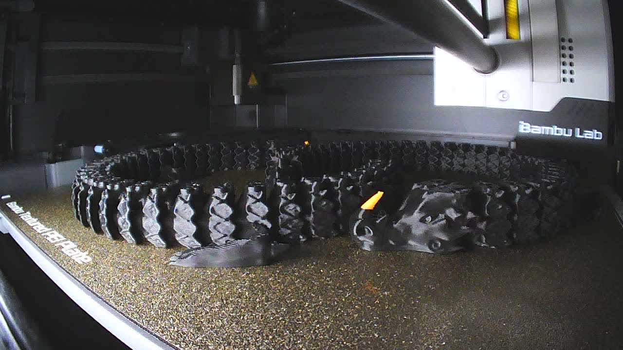 Inside a Bambu Lab P1S, a dark model printing on a gold plate labeled Bambu Textured PEI Plate. The toolhead shows the Bambu Lab name.